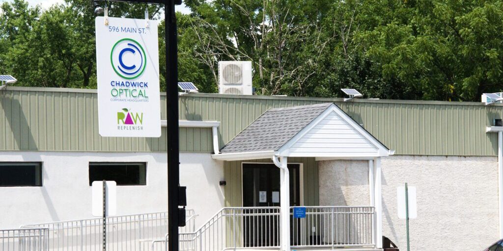 Facilities_Chadwick_Optical Exterior of Chadwick Optical corporate headquarters, showing a white building with green accents and a hanging sign displaying the company logo.