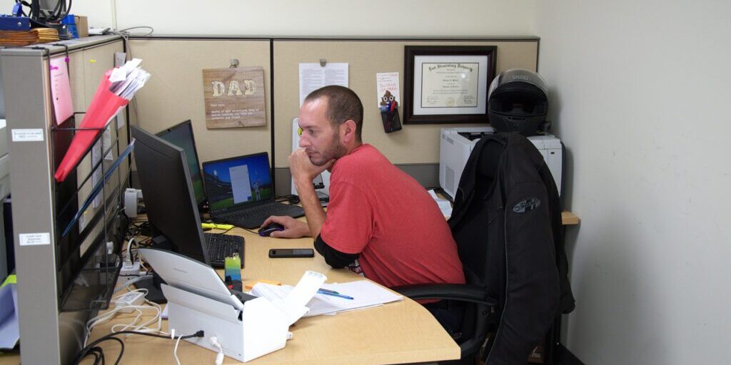 Finance_Chadwick_Optical Man in a red shirt focused on his computer at a cubicle desk, with a motorcycle helmet and framed diploma on the wall behind him.