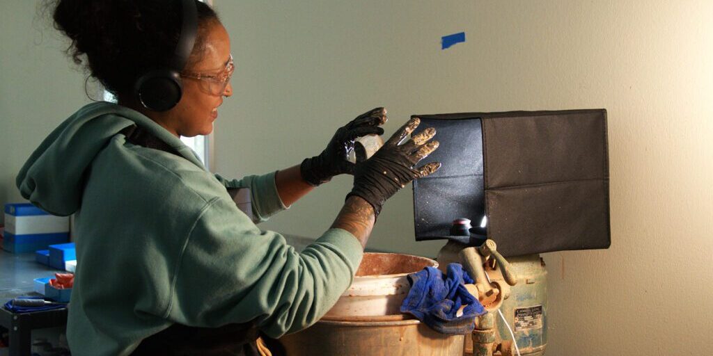 lens_casting_Chadwick_Optical Woman wearing safety goggles and gloves working on a lens casting process under focused lighting, with hands positioned over casting equipment at Chadwick Optical.