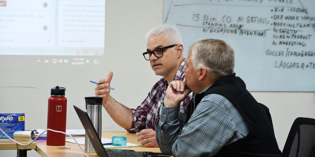 Marketing_Chadwick_Optical Two men engaged in a discussion during a meeting at a conference table, with notes visible on a whiteboard in the background.