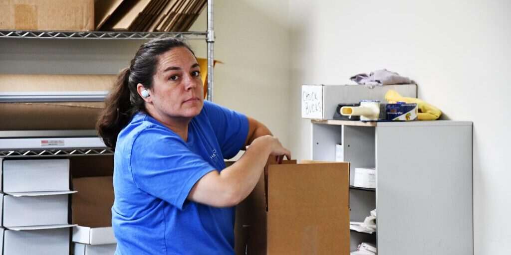 order_fulfillment2_Chadwick_Optical Woman in a blue shirt, wearing an earbud, packing a cardboard box in a workspace with shelves and packing materials.