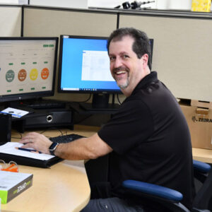 shipping_chadwick_optical Ed, Shipping Manager, smiling at his desk with dual monitors displaying shipping information.
