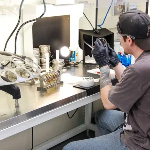 Ophthalmic-lab-technician Lab technician seated at a workbench, inspecting a lens with various optical tools and equipment around.